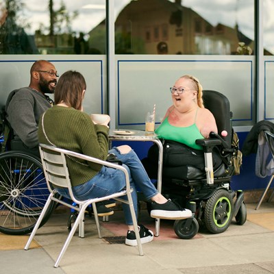 Tina is sitting with her friends laughing outside a café, they are sitting around a table. One is a lady sitting on a chair, holding a large mug. The other a gentleman in a powered wheelchair smiling.