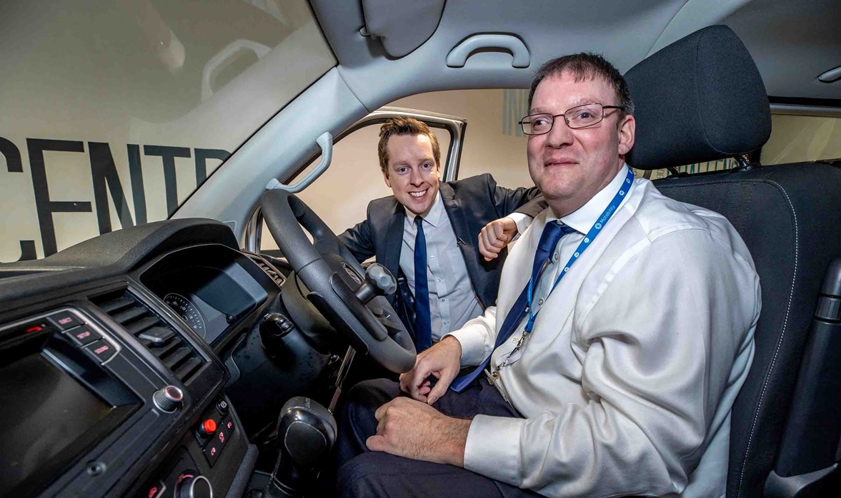 The photo shows the inside of an adapted vehicle. Sat in the front seat of the vehicle is a Motability employee smiling with his hand on knees, wearing a smart suit. Tom Pursglove MP is smiling and looking in the vehicle from outside next to the employee with his arm rested on the open right front door.