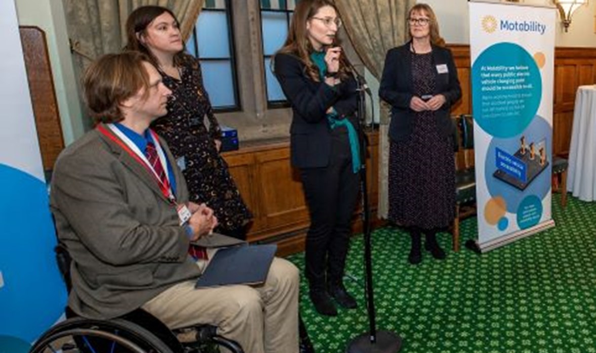 A lady is standing and talking into a microphone whilst two ladies and a man in a wheelchair listen. 