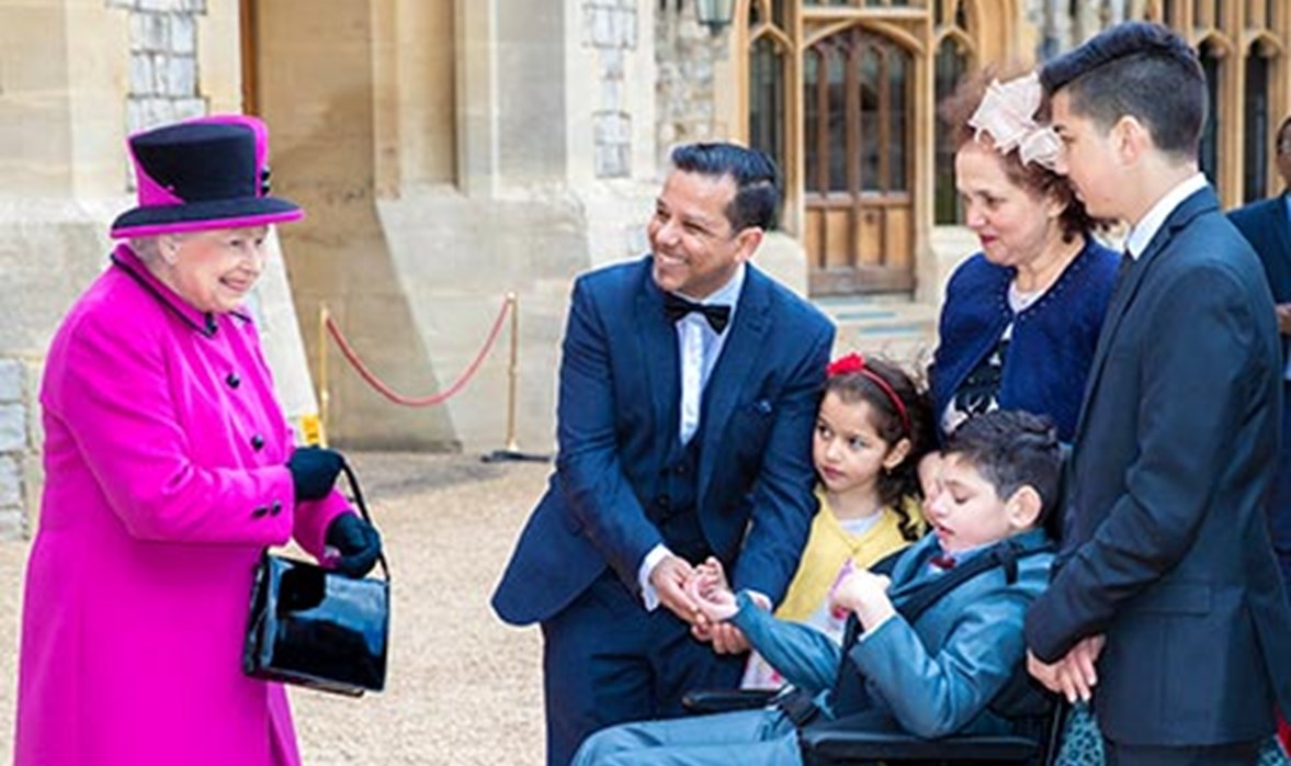 Her majesty the queen is in a pink coat and hat, she is smiling and standing next to a family. The family are smiling at her majesty.