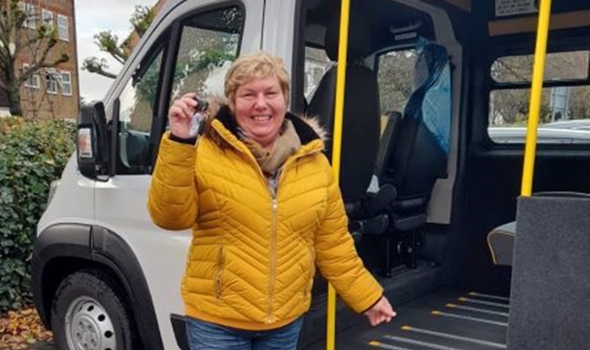 Lady smiling and proudly holding out the keys to a community transport vehicle, which is behind her. 