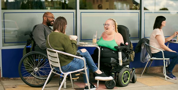 Tina is sitting with her friends laughing outside a café, they are sitting around a table. One is a lady sitting on a chair, holding a large mug. The other a gentleman in a powered wheelchair smiling.