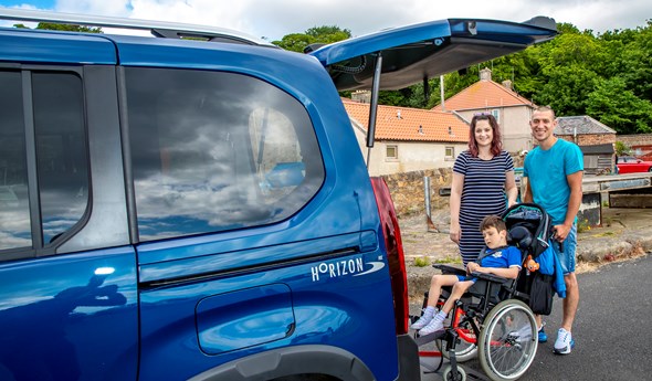 A boy in a wheelchair is in front of a women and man next to a car.