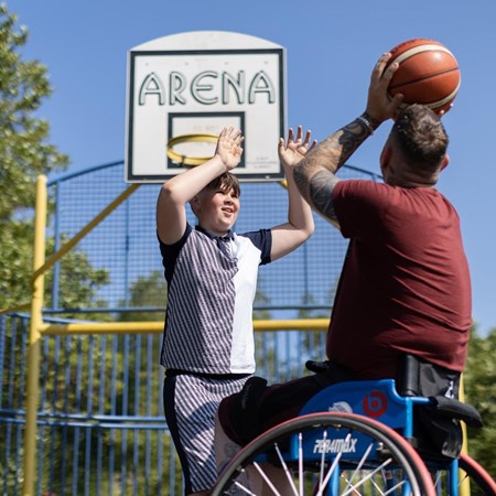 Aaron is sitting in his wheelchair on a basketball court with his son. His son is reaching for the basketball which defending the net. Aaron is throwing the ball. 