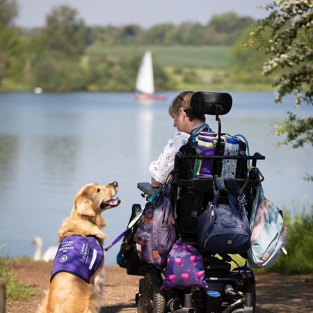 A smiling women in a wheelchair is next to a lake with a dog.