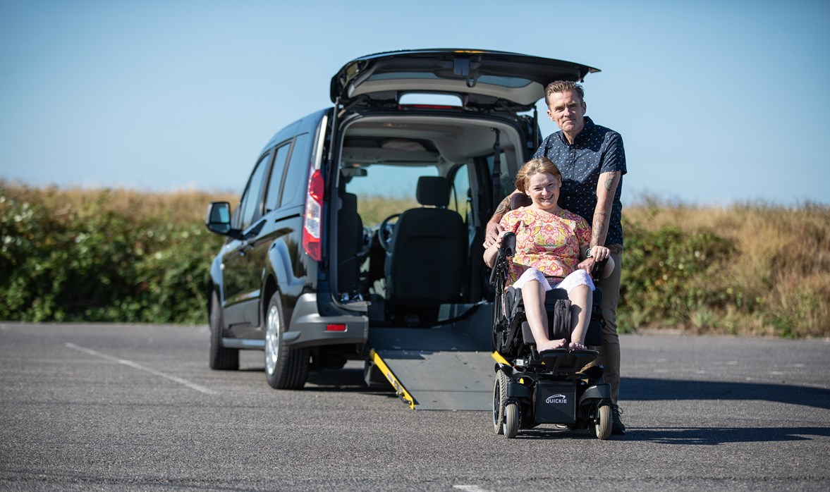 Joanne is sat in her motorised wheelchair outside at the back of her WAV, with her husbands arm around her 