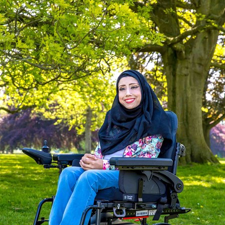Nazmin is in her powerchair, outside on a sunny day. She's wearing a dark hijab, bright shirt and a big smile