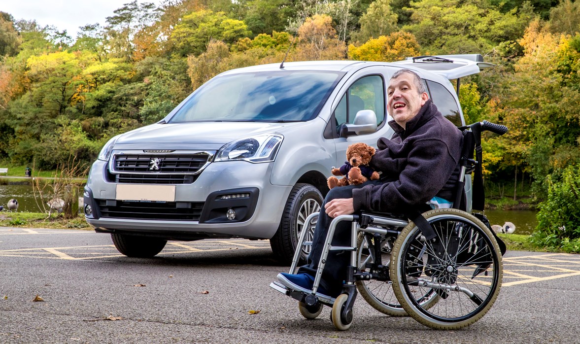 Kevin's WAV is parked up, he is outside it, in his wheelchair, with a Motability teddy bear on his lap