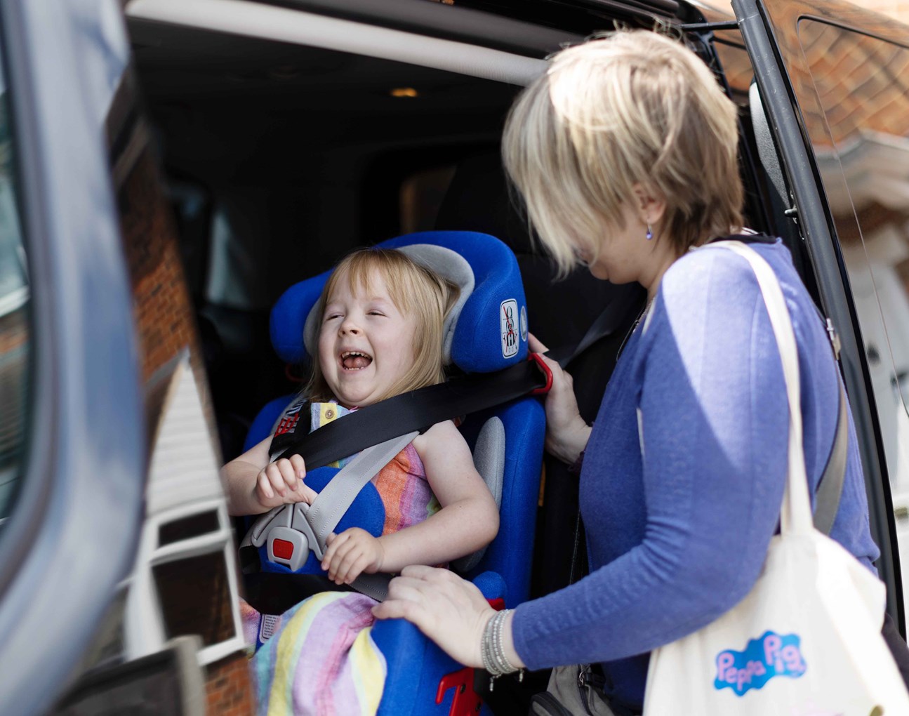Poppy's car door is slid open. Poppy has a big smile on her face. Her mum is carefully strapping her into car seat before they take a trip out for the day.