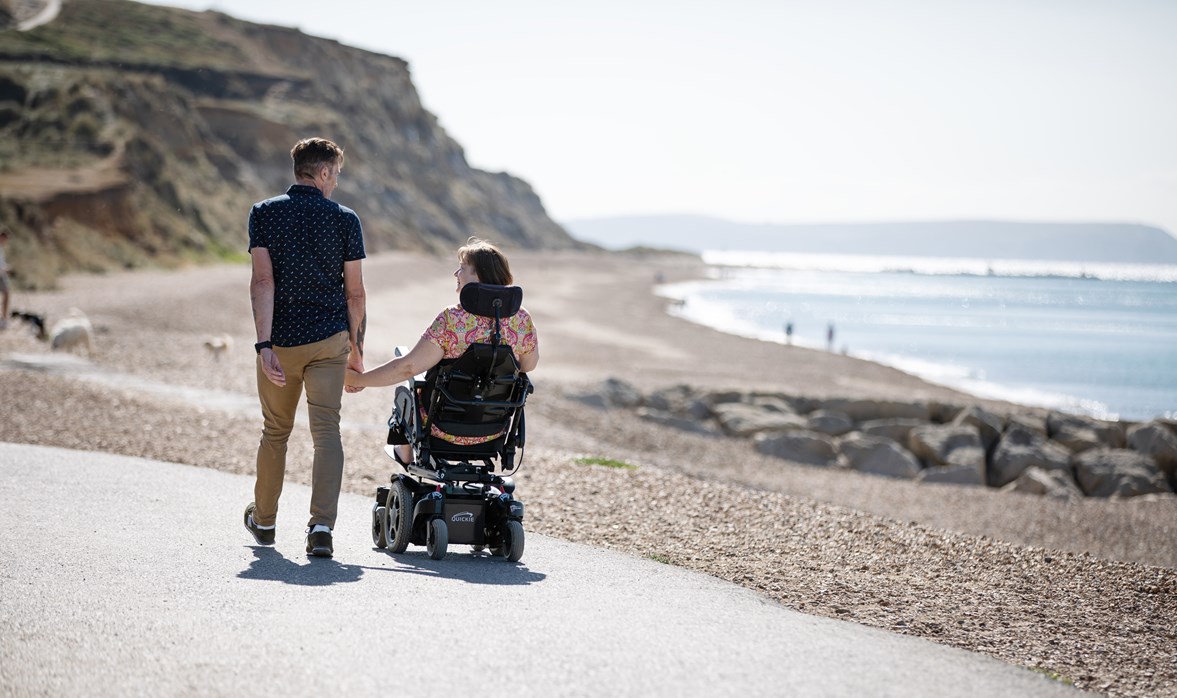 Joanne and her husband are at the beach; she is in her motorised wheelchair, they're holding hands