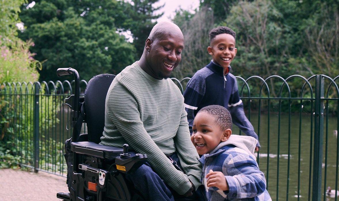 A man in a wheelchair plays with his two sons beside a lake in a park