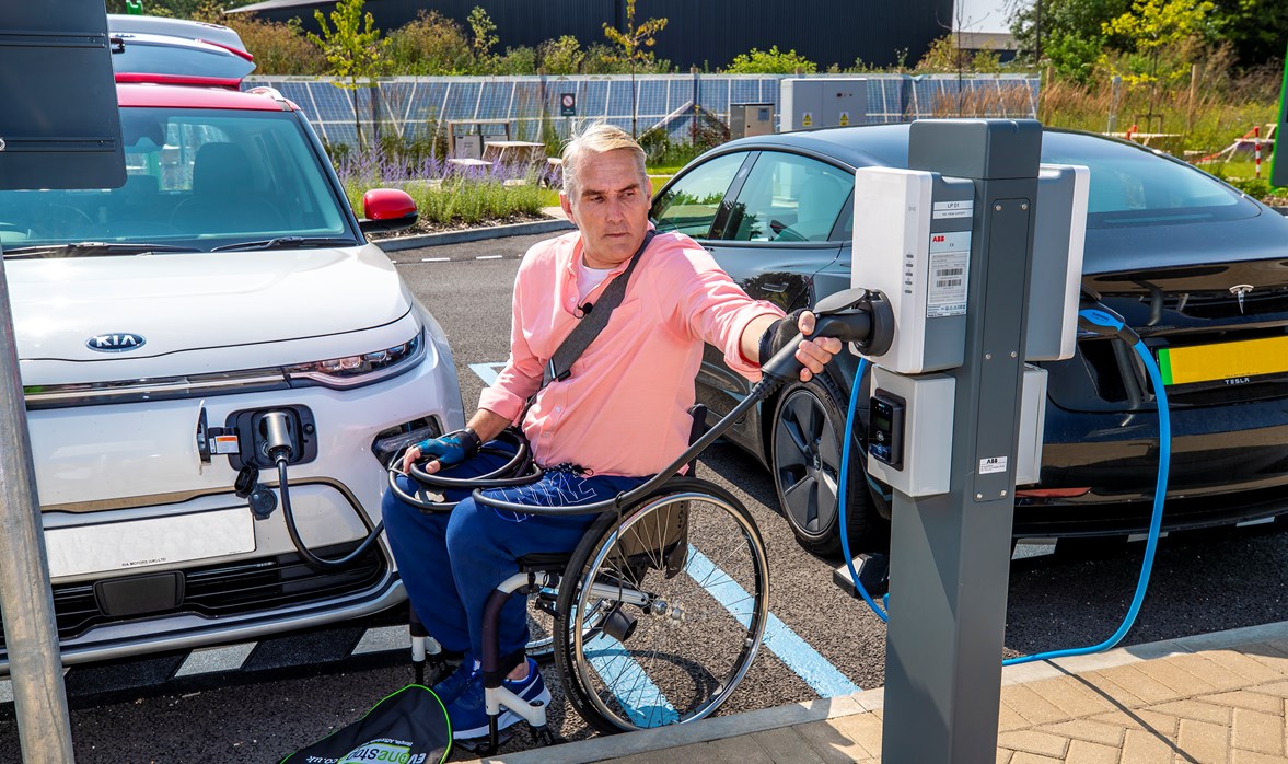 A man in a wheelchair is plugging in his electric car into an electric vehicle charging-point. 