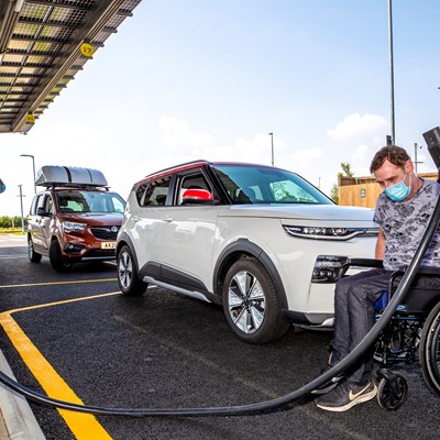 A man in a wheelchair is holding a charging plug for an electric car. The wheelchair is in front of a parked electric vehicle.