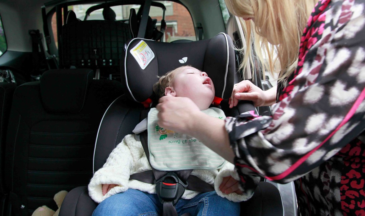 A baby boy is within a car, sitting in a car seat while his mum is securing the car seat safely. 