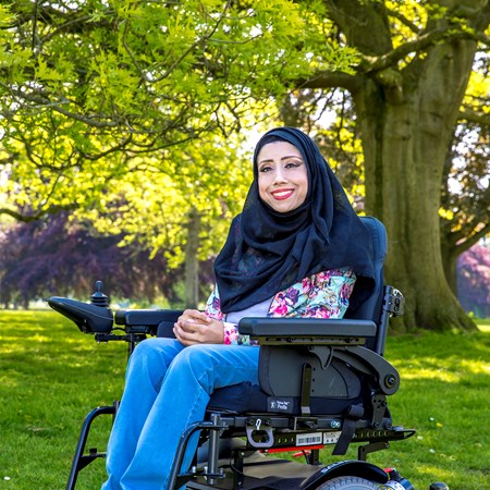 Nazmin is in her powerchair, outside on a sunny day. She's wearing a dark hijab, bright shirt and a big smile