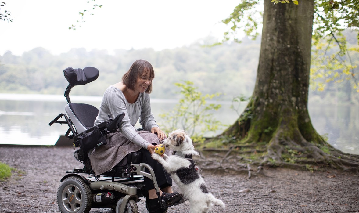 Michelle is by a lake, she sits forward in her motorised wheelchair as her dog jumps up to get the ball from her hand