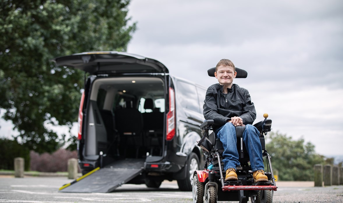 Charlie is sitting in his powerchair outside his WAV on a cloudy day