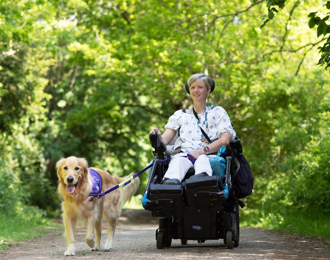 A smiling women in a wheelchair is moving through a wood with a dog. 