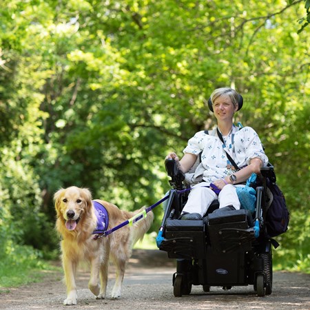 A smiling women in a wheelchair is moving through a wood with a dog. 