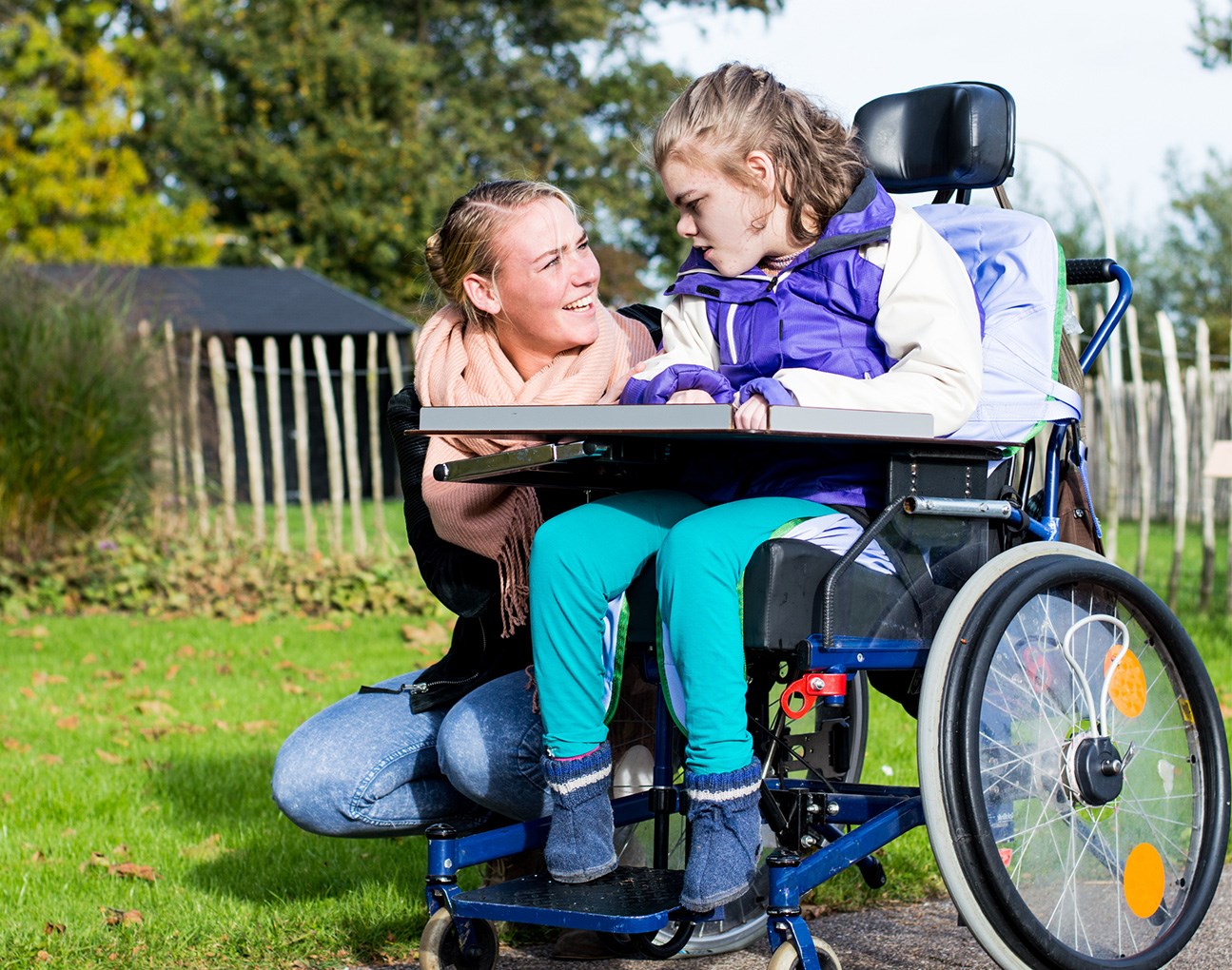 A young lady sitting in a wheelchair whilst another young lady is crouched next to her wheelchair smiling at her. 