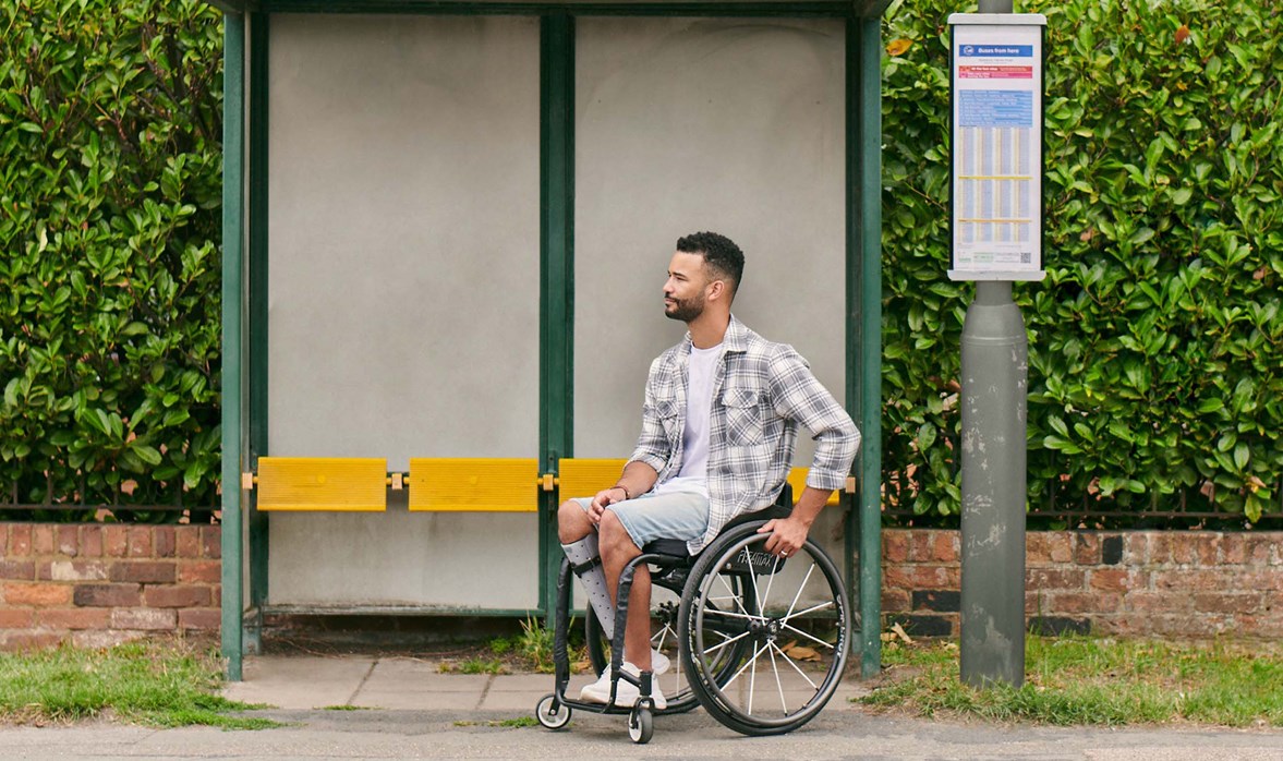 A gentleman is in a wheelchair at a bus stop waiting for a bus to arrive. 